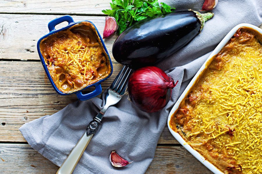 A small and large casserole filled with vegan Moussaka, on a decorative towel, with fresh red onion and eggplant.