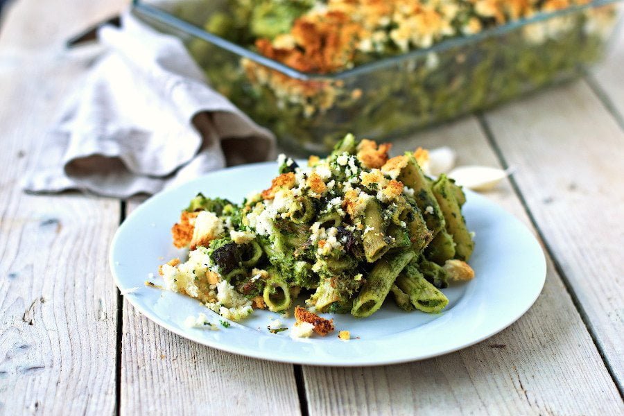 Portion of the kale pasta casserole on a white plate, rest of the casserole in the background.