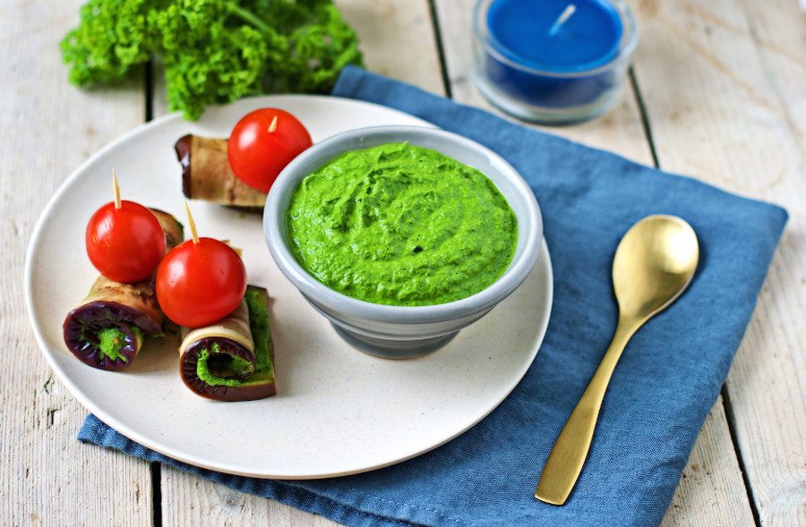 Small bowl of the Kale Pesto on a plate with zucchini rolls and tomatoes.