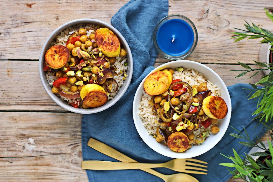 Two bowls of Vegetable Curry with Plantains on a blue cloth, with cutlery at the side.