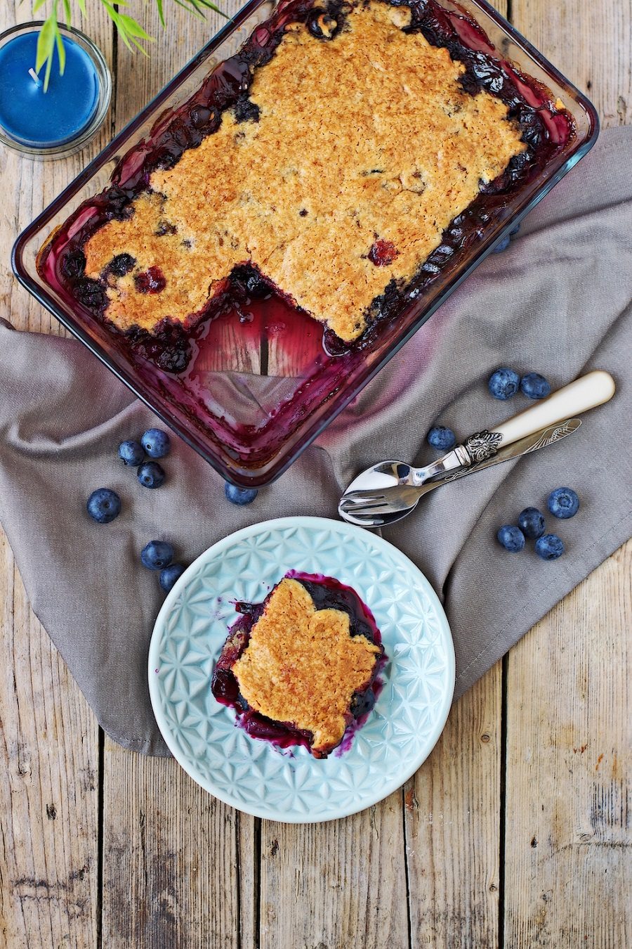 A glass casserole dish with the easy Easy Blueberry Cobbler seen from above, a portion is taken out and on a small plate.