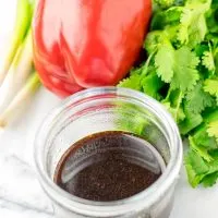 Side view of a glass jar, filled with dark sauce in front of assorted vegetables.