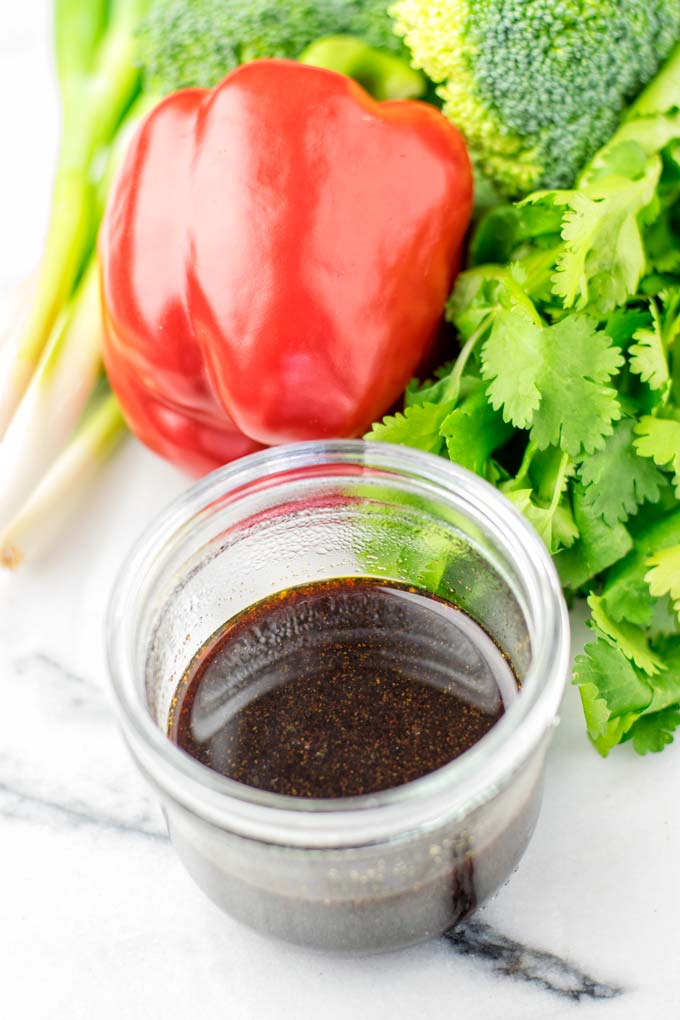 Side view of a glass jar, filled with dark sauce in front of assorted vegetables.