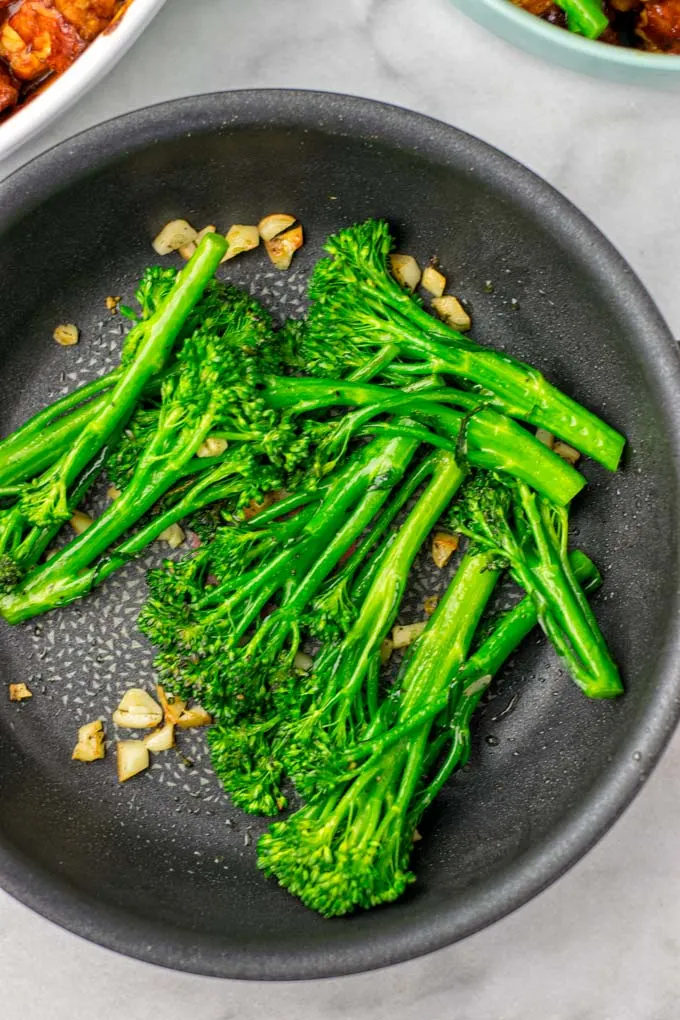 Broccoli is roasted with a touch of oil in a pan, seasoned with salt, pepper, and garlic slices.