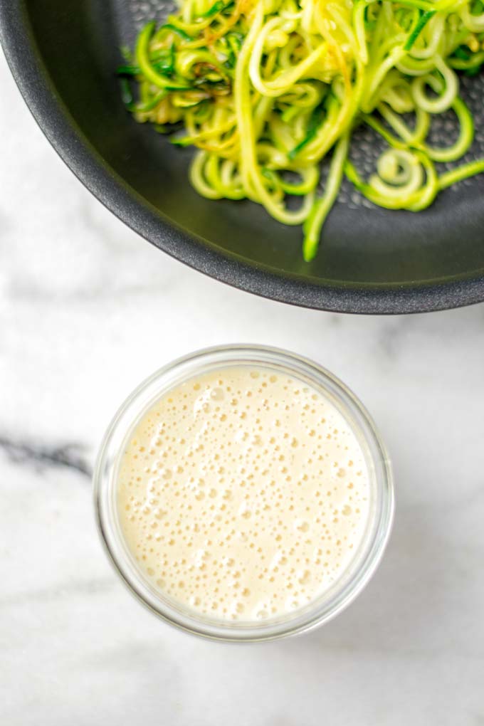 Keto Alfredo Sauce in a glass jar with a sauce pan full of zoodles in the background.