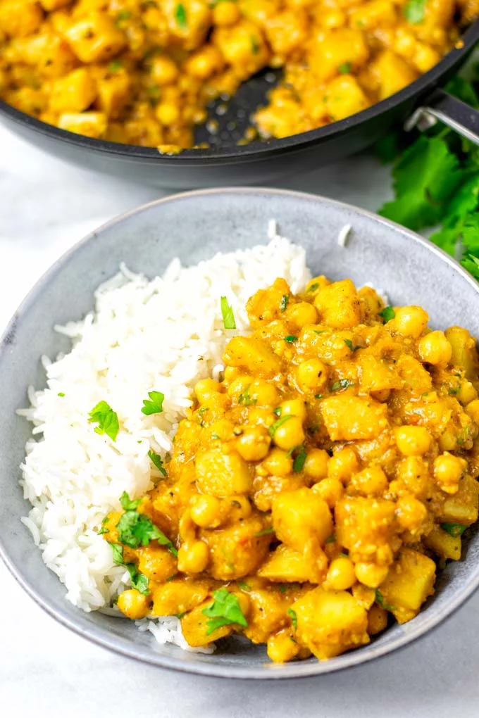 A portion of potato curry served over rice on a grey plate, with the pan in the background.