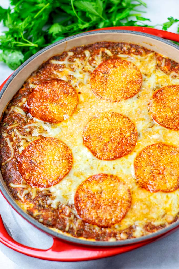 View of the baked Pizza Dip in a red casserole dish with green herbs in the background.