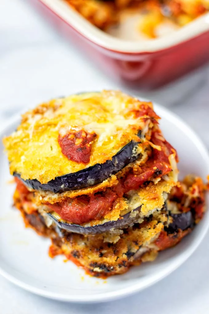 Portion of Eggplant Parmesan on a small white plate with casserole dish in the background.