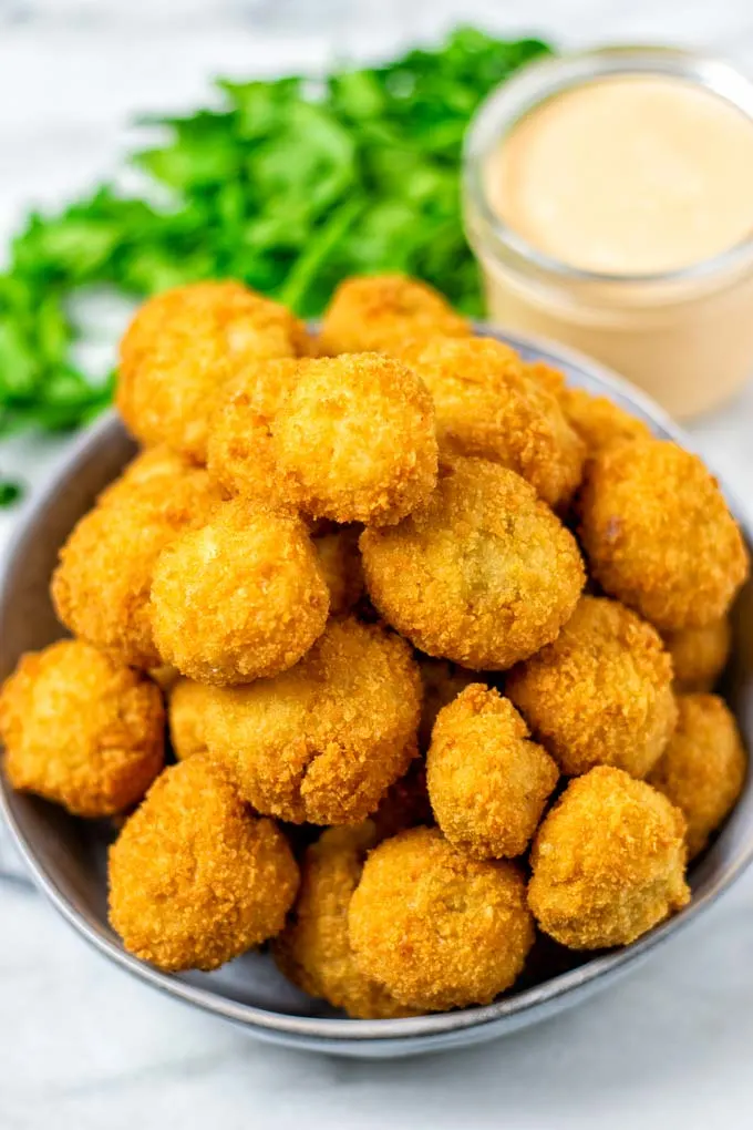 Large bowl full of ready Fried Mushrooms, with golden brown crust and a dipping sauce in the background.