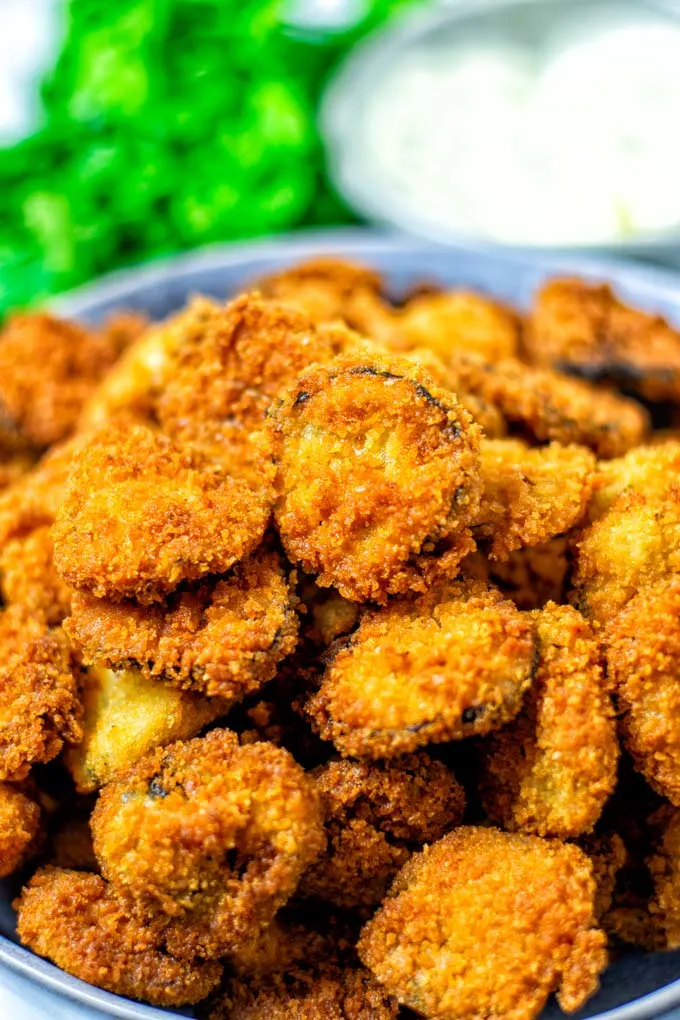 View on a batch Fried Pickles in a bowl, with some dipping sauce and fresh herbs in the background. 