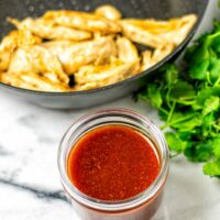 The Nashville Hot Sauce in a small glass jar is seen with a frying pan of vegan chicken and some fresh herbs in the background.