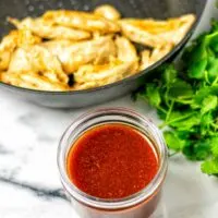 The Nashville Hot Sauce in a small glass jar is seen with a frying pan of vegan chicken and some fresh herbs in the background.