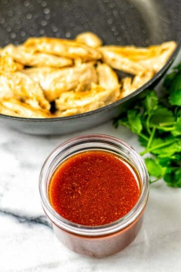 The Nashville Hot Sauce in a small glass jar is seen with a frying pan of vegan chicken and some fresh herbs in the background.