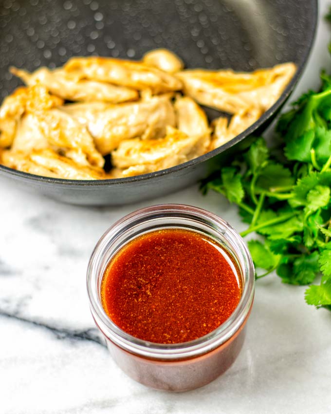 The Nashville Hot Sauce in a small glass jar is seen with a frying pan of vegan chicken and some fresh herbs in the background.