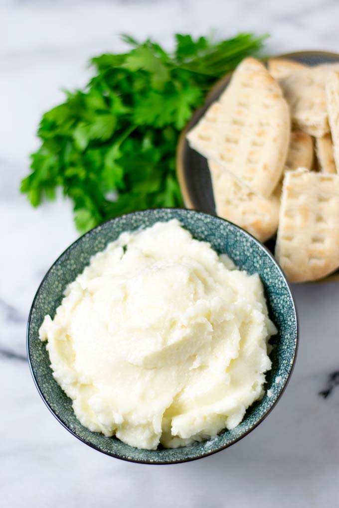 The Garlic Sauce in a serving bowl is shown with roasted pita bread and herbs in the background.