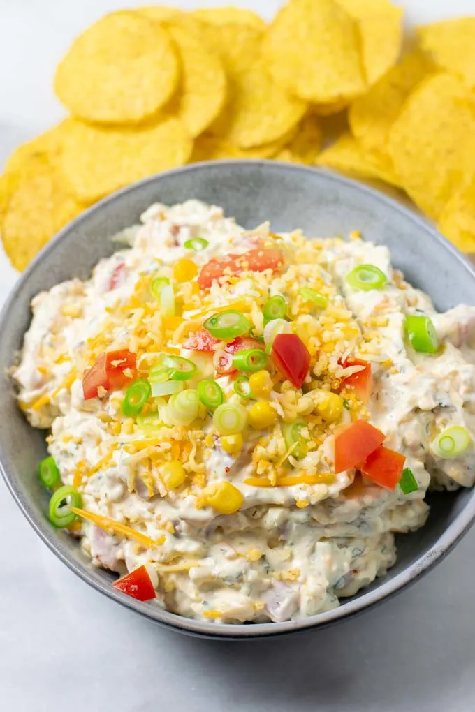 The Fiesta Ranch Dip in a serving bowl, topped with extra vegan cheese, tomatoes and scallions. Some Nacho Chips in the background.