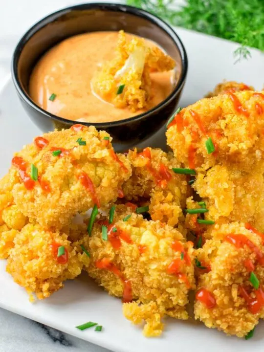 A plate with several pieces of Bang Bang Cauliflower, garnished with fresh chives and hot sauce. A small bowl with dipping sauce is also on the plate.