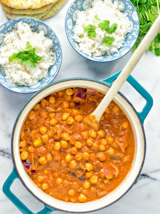 Top view on a pot with the Chickpea Tikka Masala with a wooden spoon. Also shown are two small serving bowls with rice garnished with fresh herbs and some bread in the background.