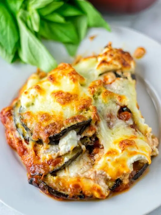 A portion of the Eggplant Lasagna is served on a plate. Layers of eggplant are clearly visible, with golden brown cheese top. Some fresh herbs in the background.