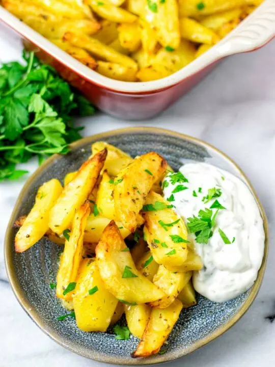 A portion of golden brown Lemon Potato wedges with a side of Tzatziki sauce is served on a blue plate, garnished with fresh herbs. More fresh herbs and a baking dish are visible in the background.