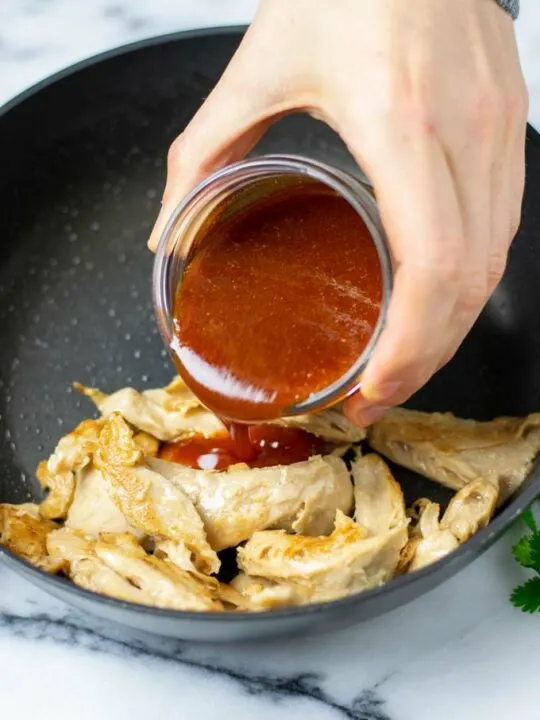 Nashville Hot Sauce is poured from a small glass jar over some fried chicken in a pan.