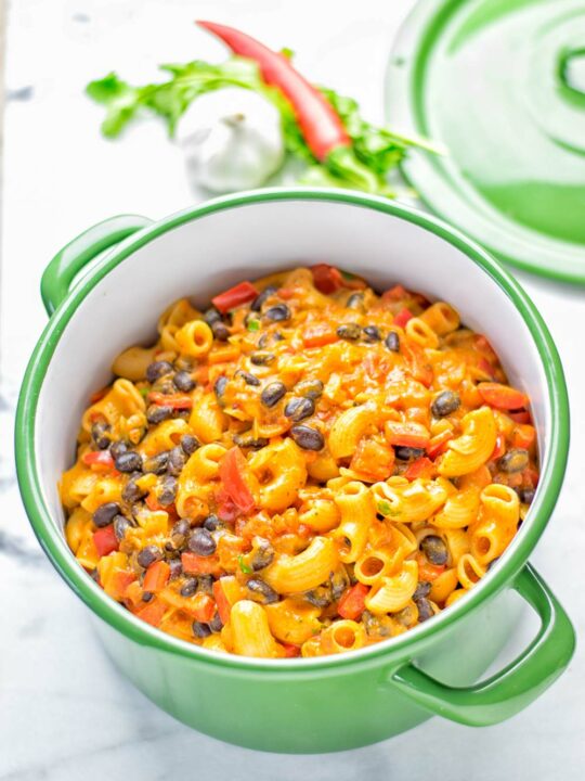 A large green saucepan with the ready Chili Mac and Cheese with some chili pepper and a head of garlic and the pot's lid in the background.