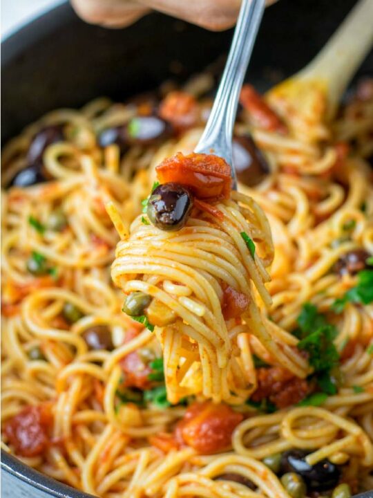 Puttanesca Sauce is served with cooked spaghetti. Closeup view of a fork with rolled up pasta and lots of sauce.