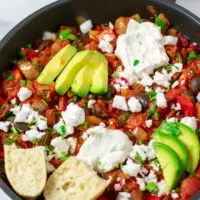 View of the finished Shakshuka in a pan, garnished with sliced avocado, vegan feta, Greek yoghurt, and some fresh bread.