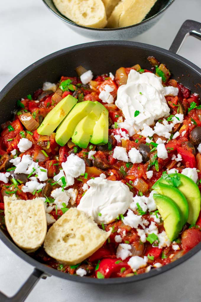 View of the finished Shakshuka in a pan, garnished with sliced avocado, vegan feta, Greek yoghurt, and some fresh bread.