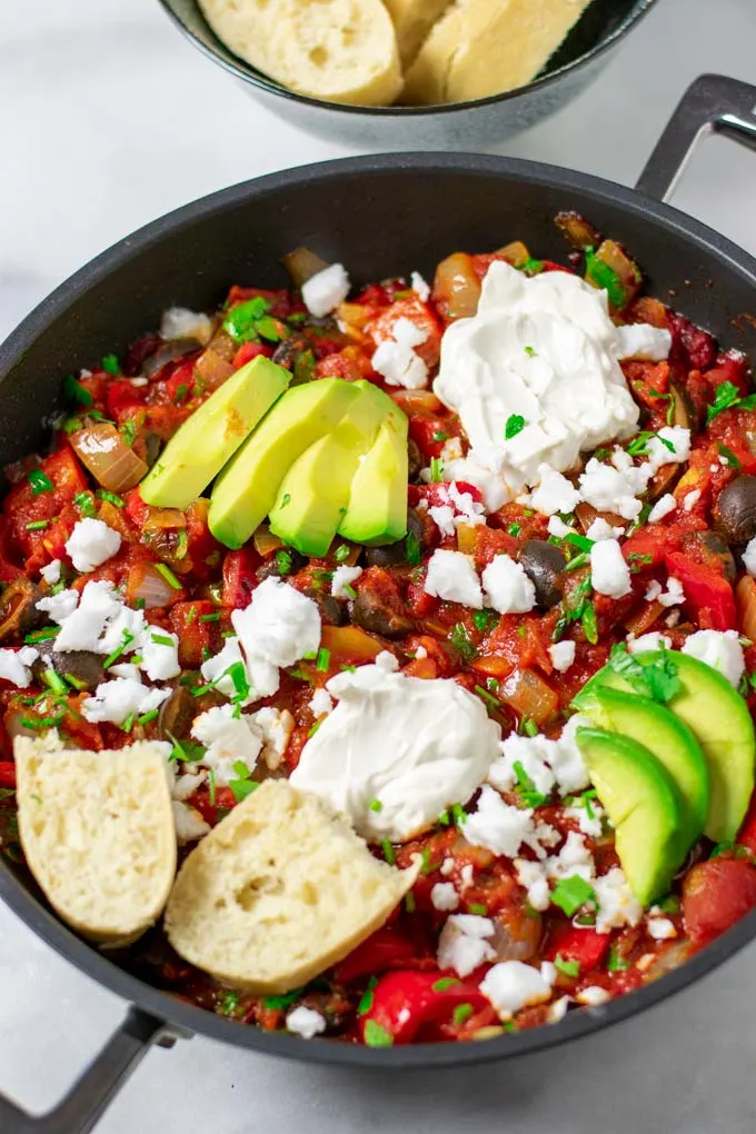 View of the finished Shakshuka in a pan, garnished with sliced avocado, vegan feta, Greek yoghurt, and some fresh bread.