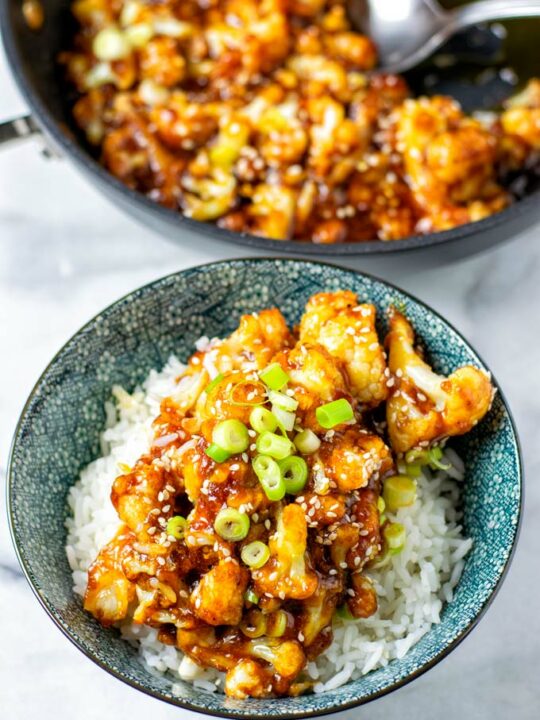 Portion of Sweet and Sour Cauliflower given over a blue bowl full of rice, with a frying pan in the background.