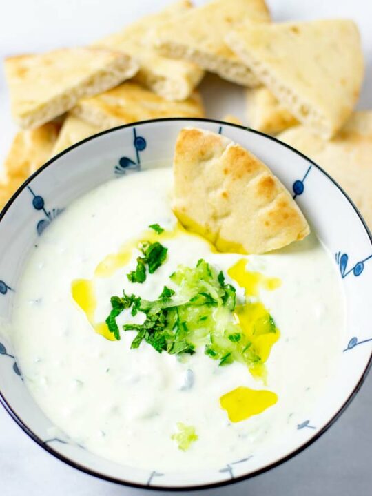 A large serving bowl with the Tzatziki sauce, garnished with extra shredded cucumber, fresh herbs. A piece of pita bread is dipped into the sauce. More slices of bread in the background.