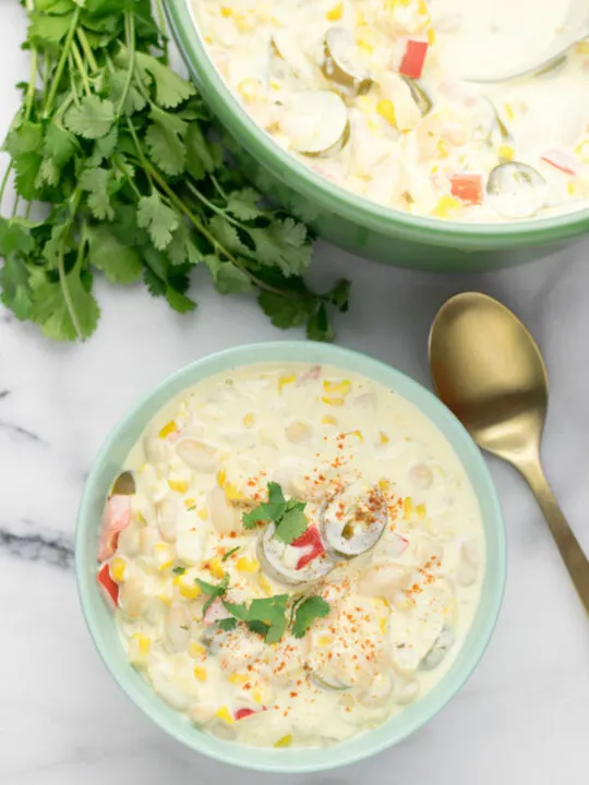 View of a portion of the creamy Vegetarian White Chili in a bowl, with a golden spoon at the side and fresh herbs. In the background is a pot with more of the chili.