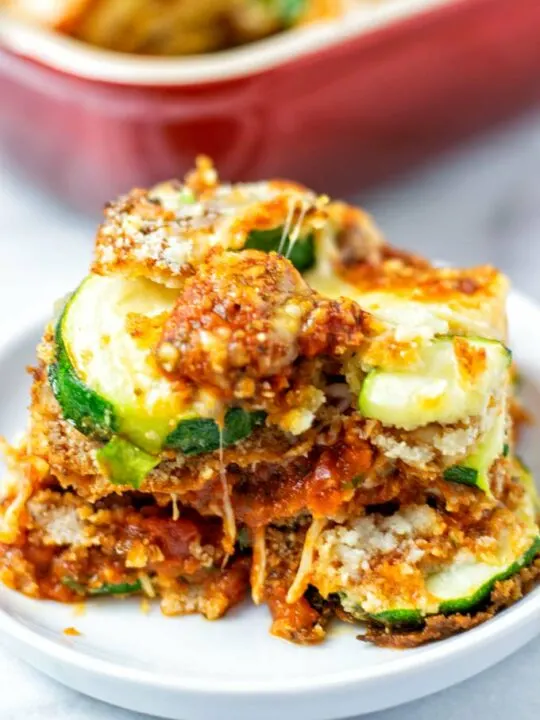 A closeup of a portion of the Zucchini Parmesan on a white plate, with a baking dish in the background. 