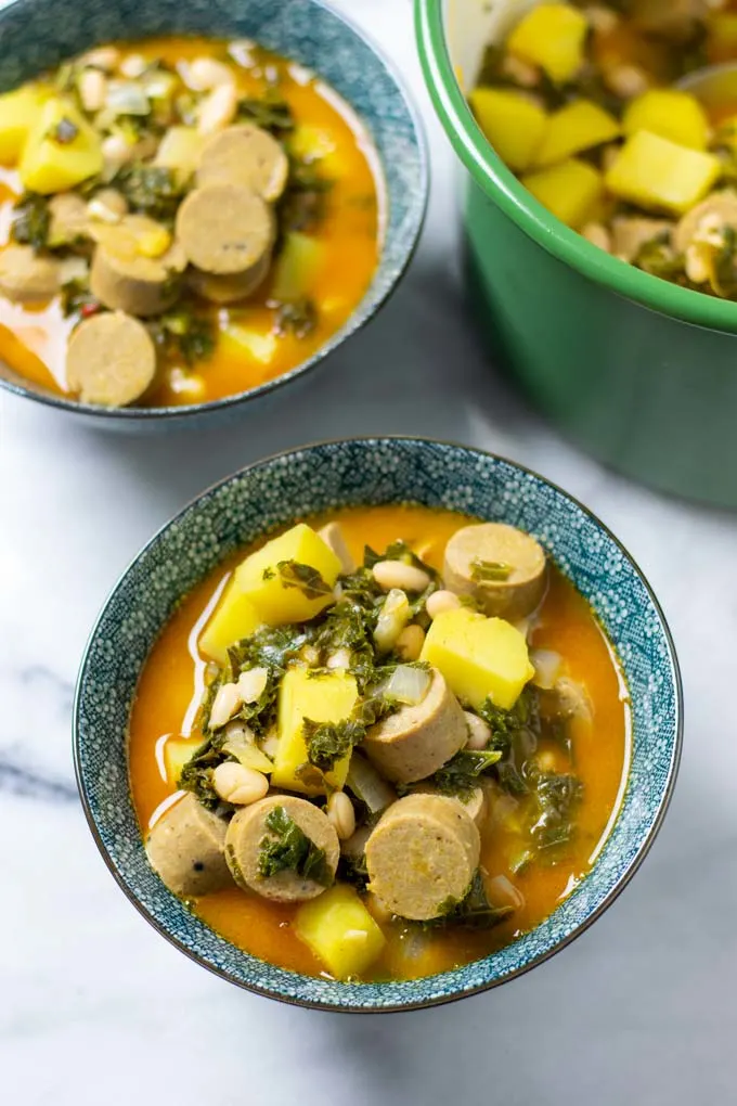 Two serving bowls with the Kale Soup on a white surface, with the soup pot in the background. 