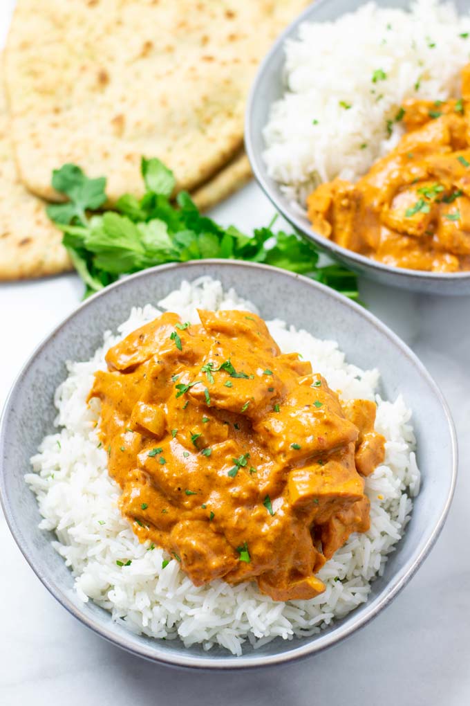 Shows two plate of Butter Chicken served with rice, some fresh cilantro and naan in the background.