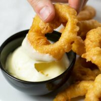 A hand is holding an onion ring and dips it into a small bowl with vegan mayo.
