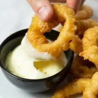 A hand is holding an onion ring and dips it into a small bowl with vegan mayo.