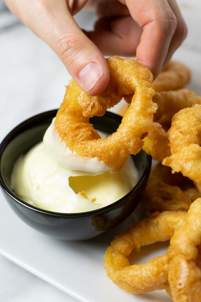 A hand is holding an onion ring and dips it into a small bowl with vegan mayo.