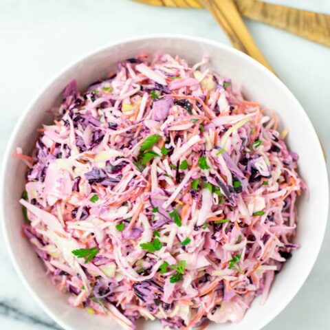 Top view on a white serving bowl with the Coleslaw, with salad cutlery in the background.