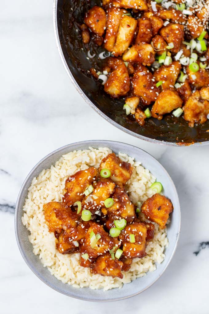 A portion of the Sweet and Sour Chicken is served on a plate with rice, garnished with scallions and sesame seeds.