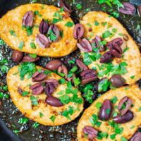 Top view of four vegan chicken breasts in a pan, with olives and fresh parsley.