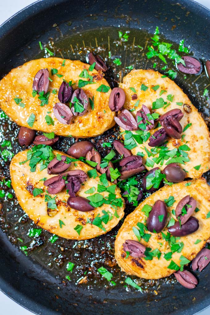 Top view of four vegan chicken breasts in a pan, with olives and fresh parsley.