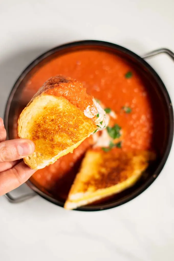 A hand lifts a sandwich dunked in tomato soup from the pot.
