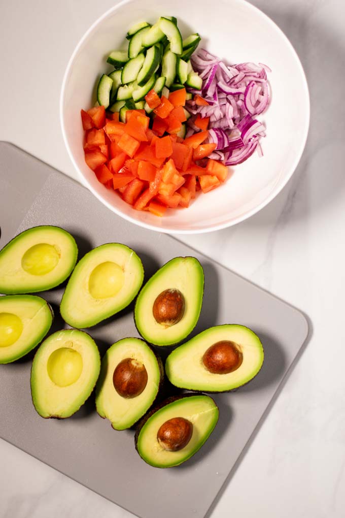 A large white mixing bowl is shown with sliced cucumber, diced tomatoes and onions, next to a cutting board with a few open avocados.