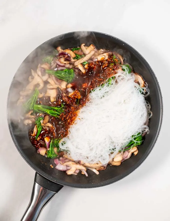 Top view of a large frying pan with broccoli, shiitake mushrooms, glass noodles, and stir Fry sauce.