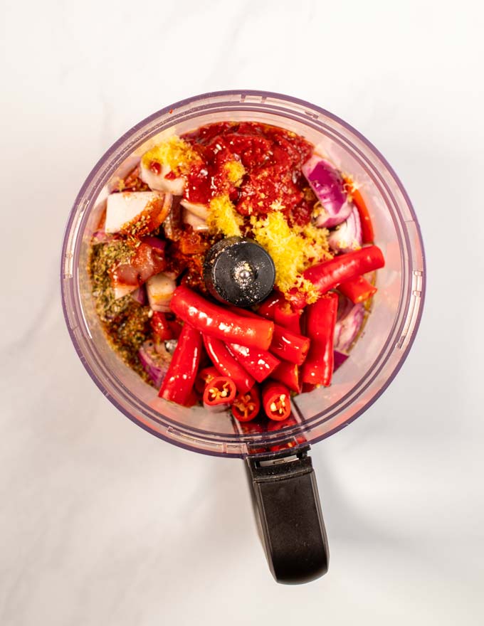 Top view into a bowl of a food processor, showing the chilis, roasted paprika, lemon zest, and garlic.