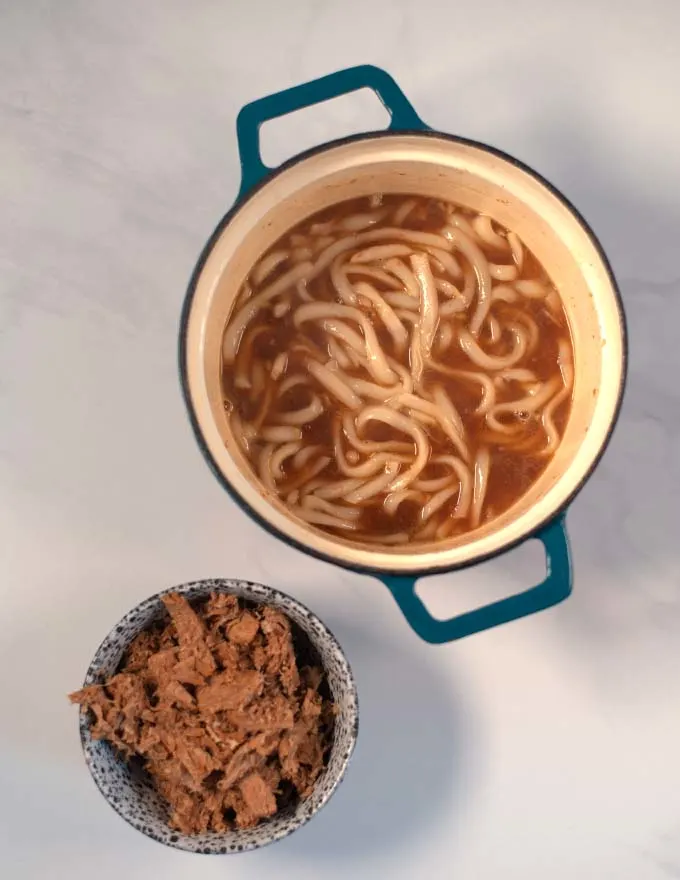 Udon noodles in a pot, with a small bowl of fried beef stripes.