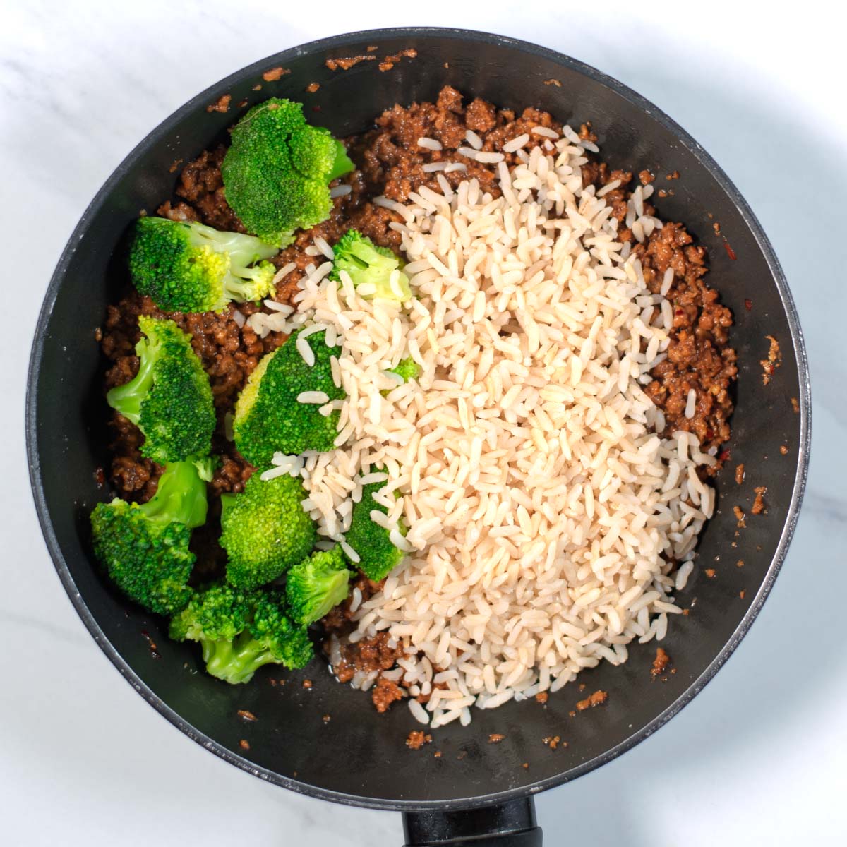 Cooked rice and broccoli are mixed with the ground beef in the pan.