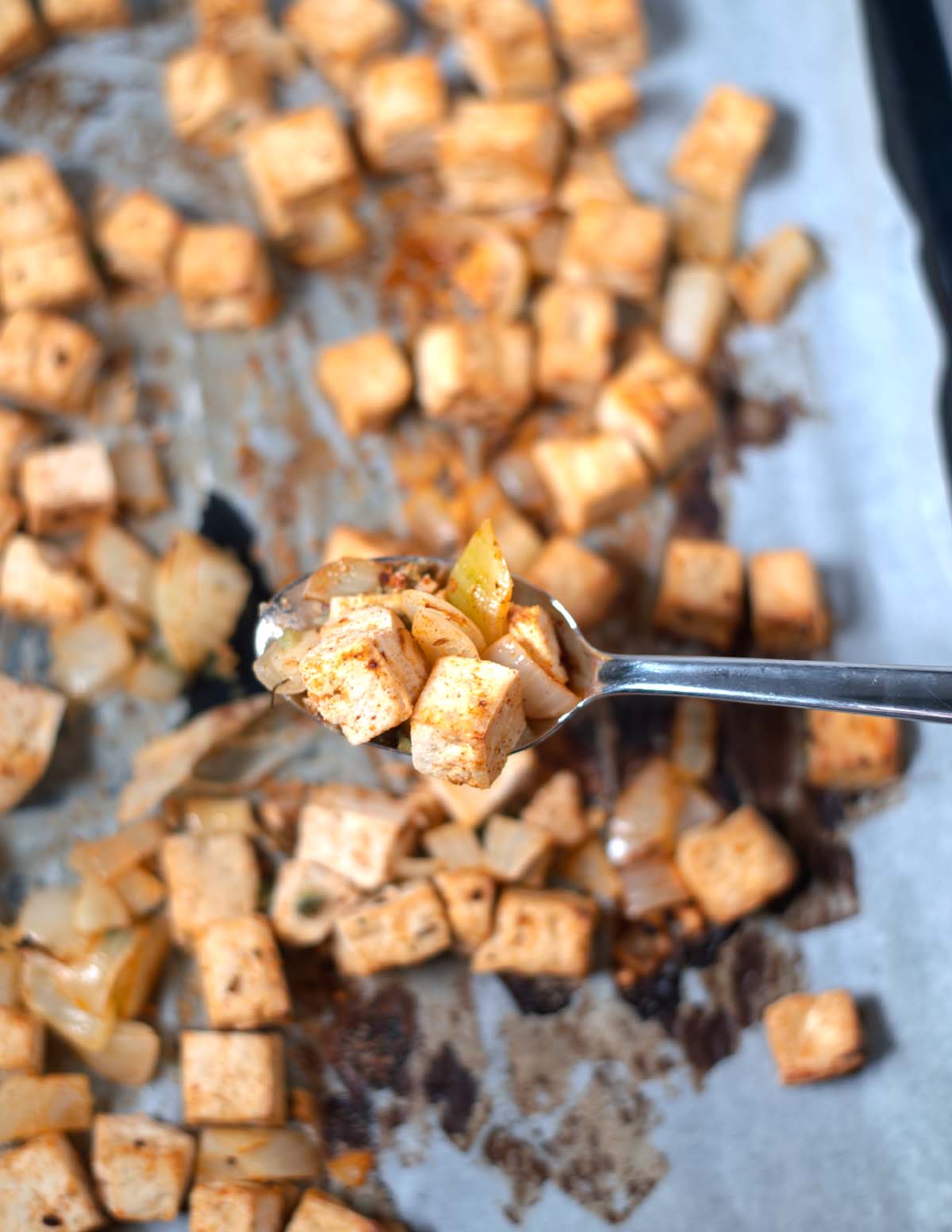 A spoon lifts roasted tofu cubes from the sheet pan.
